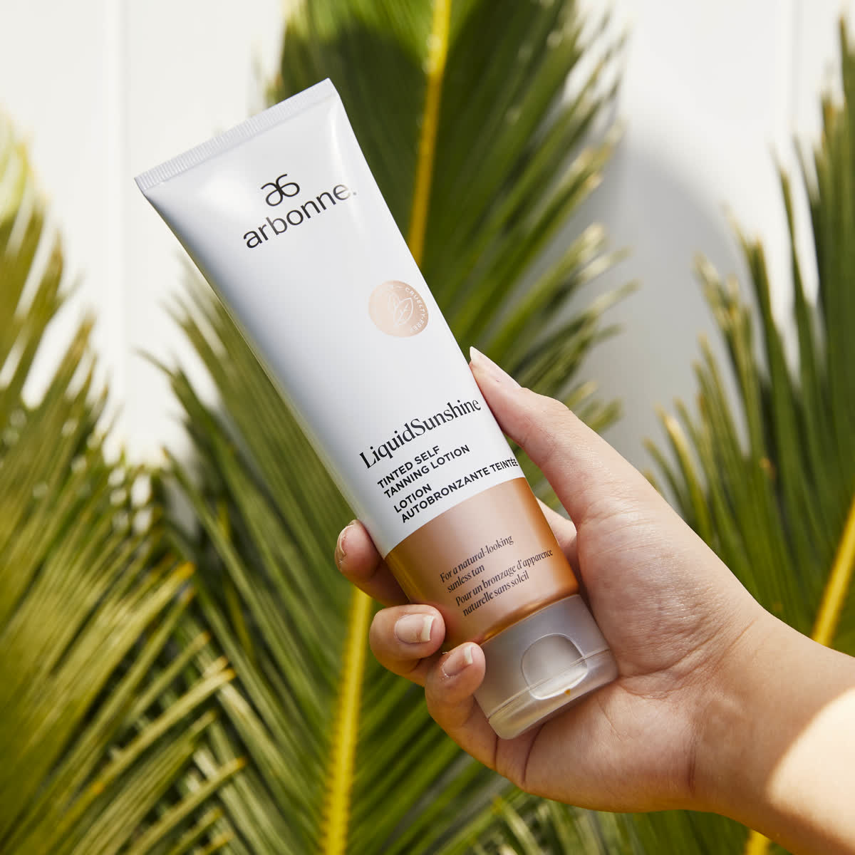 Hand holding Arbonne LiquidSunshine lotion against a palm frond